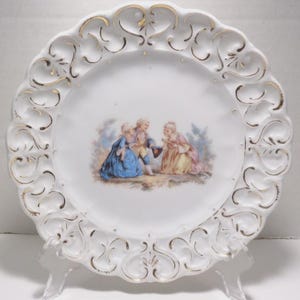 May include: White porcelain plate with a scalloped edge and gold trim. The plate features a central image of three figures in period clothing. The plate is displayed on a clear plastic stand, showcasing its decorative design.