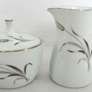 May include: A white porcelain sugar bowl and creamer set with a silver wheat design. The sugar bowl has a lid and the creamer has a handle.