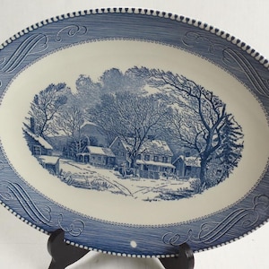 May include: An oval ceramic platter featuring a blue and white transferware pattern. The center depicts a winter landscape with houses, trees, and snow. The wide border has a textured blue finish with decorative scrollwork and a beaded edge.