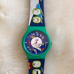 May include: A vintage Snoopy watch with a green case and a blue strap. The watch face features Snoopy and other Peanuts characters in an underwater scene. The strap has a whale and more Peanuts characters.