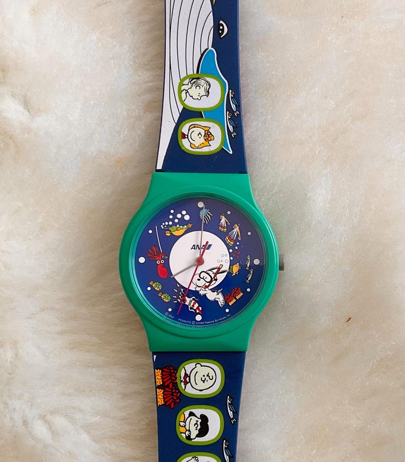 May include: A vintage Snoopy watch with a green case and a blue strap. The watch face features Snoopy and other Peanuts characters in an underwater scene. The strap has a whale and more Peanuts characters.