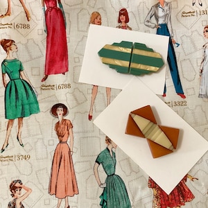 May include: Two decorative hair clips on white cards. The top clip is a geometric, elongated shape in alternating horizontal stripes of matte teal and pearlescent cream. The bottom clip is a rust-orange angular piece with a central cream-colored inlay.
