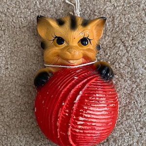 May include: A vintage ceramic cat figurine with orange and black markings, holding a large red ball of yarn. The cat has black paws and big eyes. A white string is attached at the top for hanging.