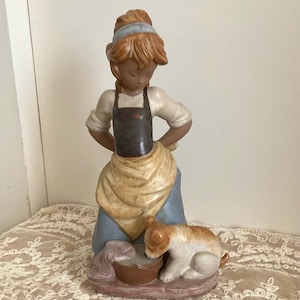 May include: A porcelain figurine depicting a person with brown hair wearing a white shirt, a dark brown apron, and a blue skirt, standing over a small brown basin. A small white and brown dog sits beside the basin on a base with a pinkish hue.
