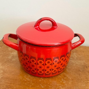 May include: A red enamel pot with a matching lid. The pot features a floral pattern in shades of orange and brown. It has two side handles and a loop handle on the lid. The pot is sitting on a wooden surface.