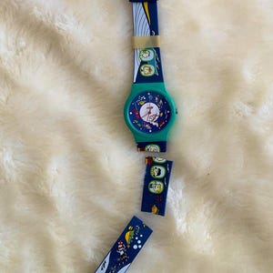 May include: A vintage wristwatch with a teal face and a blue strap featuring a whimsical design. The watch has a teal case and a blue strap with colorful illustrations. The watch is lying on a white, fluffy surface.