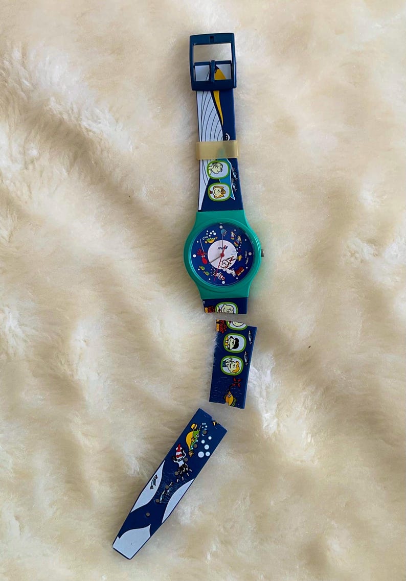 May include: A vintage wristwatch with a teal face and a blue strap featuring a whimsical design. The watch has a teal case and a blue strap with colorful illustrations. The watch is lying on a white, fluffy surface.