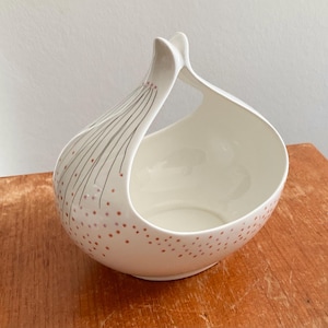 May include: White ceramic decorative bowl with a unique handle. The bowl features a rounded base and a handle that curves upwards. It is decorated with small red dots and thin black lines with red dots.