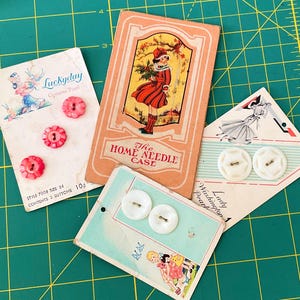 May include: Three vintage cardboard button cards and one rectangular needle case. One card features three pink floral-shaped buttons, another holds two white buttons, and the third displays two white buttons. The needle case shows a child illustration.