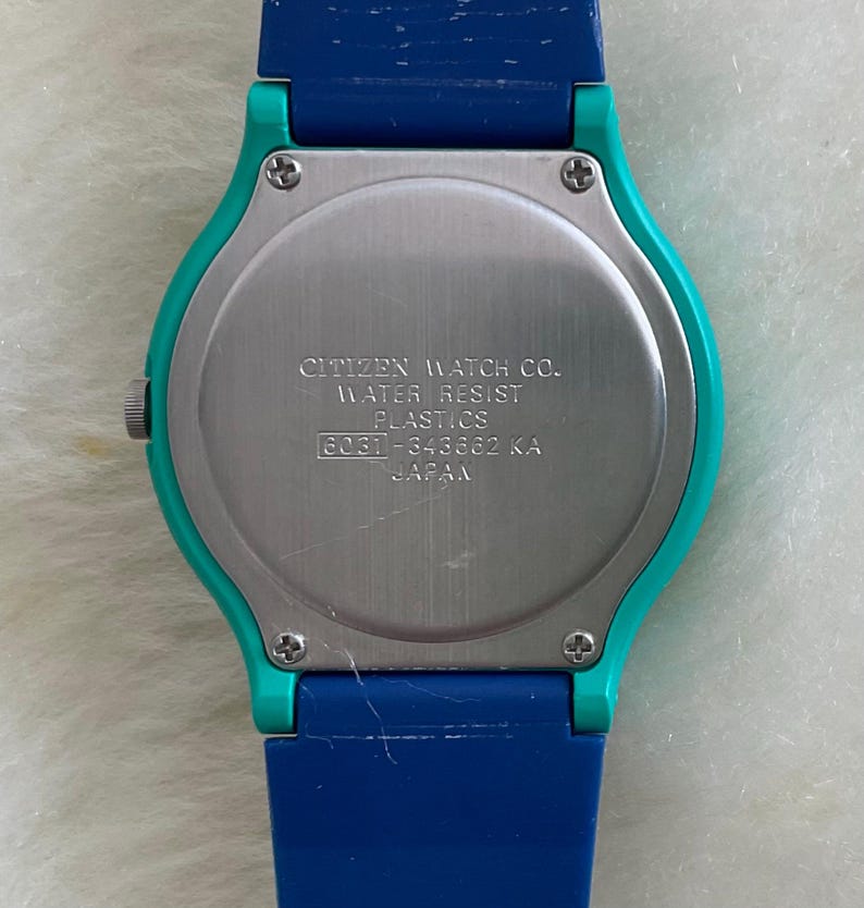 May include: A close-up of the back of a Citizen watch with a blue and teal strap. The watch face is silver with the text "CITIZEN WATCH CO. WATER RESIST PLASTICS 8031-343662 KA JAPAN".
