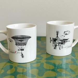 May include: Two white ceramic mugs featuring black ink illustrations of vintage-style flying machines. Each mug displays a detailed airship or aircraft design with the text Virgin Atlantic printed in small red and black lettering on the side.