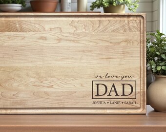 May include: A rectangular light-colored wooden cutting board with a juice groove along the perimeter. The board features an engraved design that reads "we love you DAD" inside a rectangular border, with the names "JOSHUA", "LANIE", and "SARAH" below.