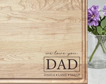 May include: A light-colored wooden cutting board with a juice groove. The board is engraved with the words "we love you DAD" and the names Joshua, Lanie, and Sarah. A vase of purple flowers is in the background.