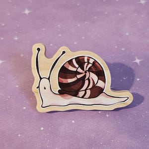 May include: A wooden snail pin with a brown and white striped shell. The snail is painted in a cartoon style and has a white body.