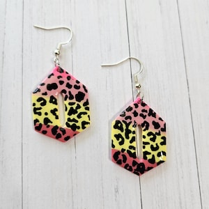 May include: A pair of dangle earrings with a geometric shape and a gradient of pink and yellow. The earrings feature a black leopard print pattern and silver-colored ear wires.