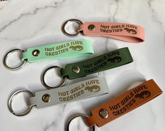 May include: A collection of five keychains in various colors: pink, mint green, dark green, gray, and orange. Each keychain has a silver ring and a leather strap with the text "HOT GIRLS HAVE CRESTIES" and a lizard graphic.