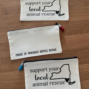 May include: Three rectangular off-white canvas zip pouches. Two pouches feature the black text 'support your local animal rescue' with a map of New York and a heart. One pouch features the black text 'HOUSE OF PANCAKES REPTILE RESCUE'.