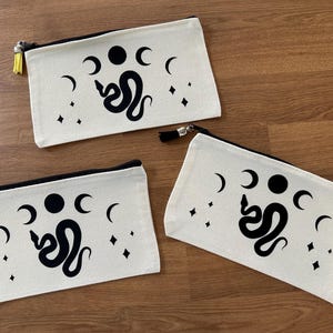 Pode incluir: Três estojos de lona com fecho de correr em tom off-white, com uma impressão gráfica preta de uma cobra rodeada por fases lunares e estrelas de quatro pontas. Cada estojo tem um fecho preto e uma borla decorativa amarela, roxa ou preta.