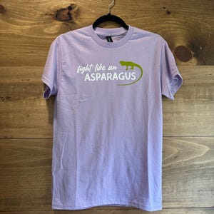 May include: A light purple short-sleeve t-shirt featuring a green lizard graphic and the white text "fight like an ASPARAGUS" printed on the chest. The shirt has a crew neckline and is displayed on a black hanger against a wooden background.