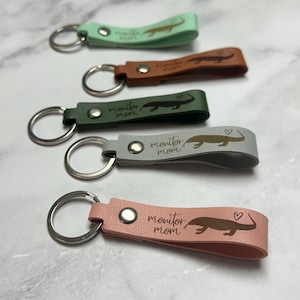 Op de afbeelding: Een verzameling van vijf leren sleutelhangers in verschillende kleuren: mintgroen, bruin, donkergroen, grijs en roze. Elke sleutelhanger heeft een hagedisafbeelding en de tekst "monitor mom".