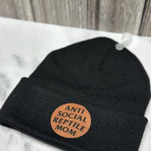 Peut inclure: Bonnet en tricot noir avec un écusson circulaire marron. L'écusson porte l'inscription "ANTI SOCIAL REPTILE MOM". Le bonnet est en tricot côtelé doux.