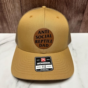 Peut inclure: Casquette de camionneur beige avec un dos en filet marron et une visière incurvée. Le chapeau comporte un écusson circulaire avec le texte "ANTI SOCIAL REPTILE DAD". Une étiquette noire et blanche est attachée à la visière.