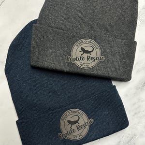 Peut inclure: Deux bonnets en tricot, un gris anthracite et un bleu marine. Chaque bonnet présente un écusson circulaire avec le texte "House of Pancakes Reptile Rescue NY-PA" et une silhouette de lézard.