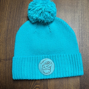Peut inclure: Un bonnet en tricot turquoise avec un pompon assorti. Le chapeau présente un écusson circulaire avec une illustration de lézard et le mot "hoppy" dans une police manuscrite.