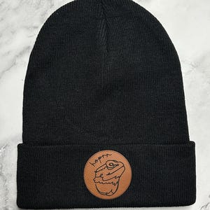 Peut inclure: Bonnet noir avec un écusson en cuir marron. L'écusson présente une illustration de lézard et le mot "hopin". Le bonnet est en maille.
