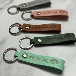 Può includere: Una collezione di portachiavi in vari colori: grigio, rosa, marrone, verde e verde menta. Ogni portachiavi presenta un cinturino in pelle con la scritta "attention hog" e un'illustrazione di un serpente, attaccato a un portachiavi argentato.