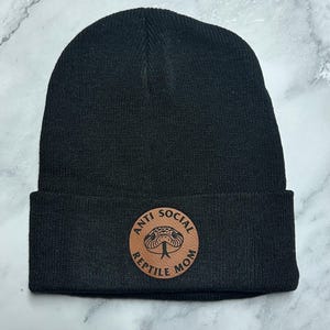 Peut inclure: Bonnet noir avec un écusson en cuir marron. L'écusson présente une illustration de serpent et le texte "ANTI SOCIAL REPTILE MOM". Le chapeau est fait d'une matière tricotée douce.