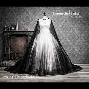 May include: A white wedding dress with a black tulle overlay and a long train. The dress has a sweetheart neckline and a fitted bodice. The tulle overlay is sheer and creates a dramatic effect. The dress is displayed on a mannequin in front of a dark patterned background.