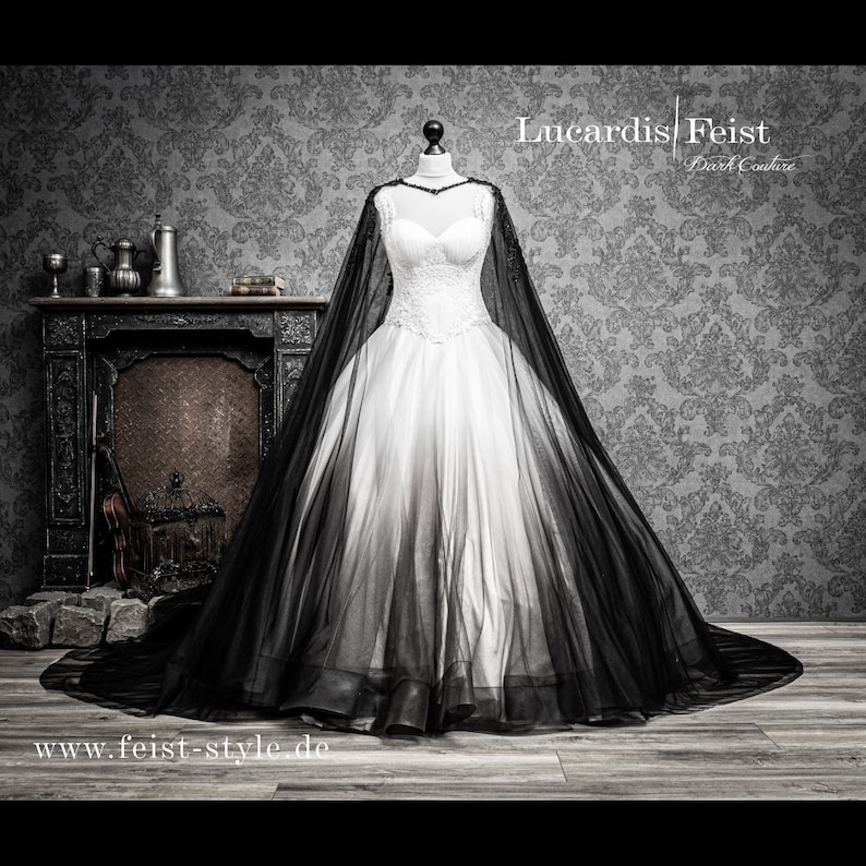 May include: A white wedding dress with a black tulle overlay and a long train. The dress has a sweetheart neckline and a fitted bodice. The tulle overlay is sheer and creates a dramatic effect. The dress is displayed on a mannequin in front of a dark patterned background.