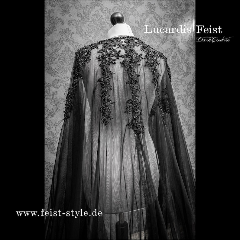May include: Black sheer lace cape with delicate floral embroidery. The cape is draped over a dress form and has a flowing silhouette. The text "Lucardis Feist Dark Couture" and "www.feist-style.de" is visible on the image.