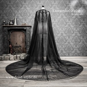 May include: A black sheer cape with lace detailing and a long train. The cape is displayed on a mannequin against a gray patterned background. The text "Lucardis Feist Dark Couture" and "www.feist-style.de" are visible in the image.