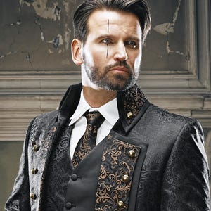 May include: A black brocade jacket featuring a dark floral pattern, a high collar, and gold-toned buttons. The jacket is worn over a white collared shirt, a black vest, and a matching black and gold patterned necktie with a dark floral design.