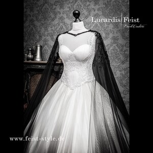May include: A white wedding dress with a lace bodice and a black tulle cape. The dress has a full skirt and is worn on a mannequin. The text "Lucardis Feist Dark Couture" and "www.feist-style.de" are visible on the image.