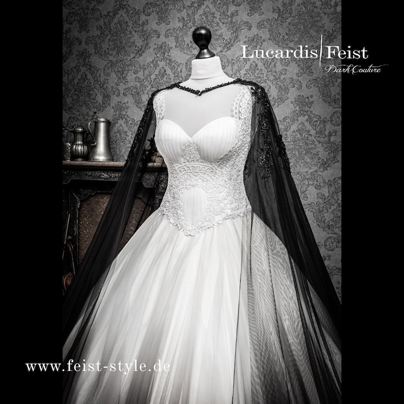 May include: A white wedding dress with a lace bodice and a black tulle cape. The dress has a full skirt and is worn on a mannequin. The text "Lucardis Feist Dark Couture" and "www.feist-style.de" are visible on the image.