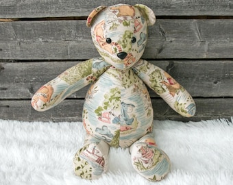 May include: A stuffed teddy bear with a patterned fabric featuring illustrations in shades of beige, blue, green, and orange. The bear has black button eyes and a black nose. The bear is sitting on a white fluffy surface.