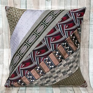 May include: A square throw pillow constructed from diagonal strips of various patterned neckties. The fabric features a mix of geometric, abstract, and repeating motifs in shades of burgundy, silver, brown, olive green, and black with a satin finish.
