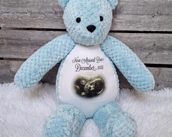May include: A light blue plush teddy bear with a textured, soft fabric finish. The bear features a white circular patch on its chest printed with a black and white ultrasound image and the text New Arrival Due December 2023 in black script.