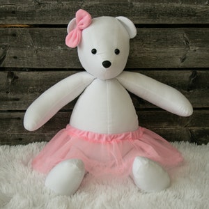 May include: A white plush teddy bear wearing a pink tutu and bow. The bear has black button eyes and a black nose. The background is a rustic wooden wall and a white fluffy rug.