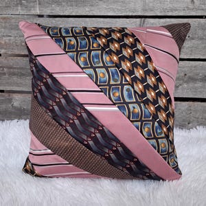 May include: A square throw pillow constructed from diagonal strips of various neckties. The design features alternating bands of solid dusty pink, dark blue with gold circular patterns, black with geometric shapes, and textured brown fabric.