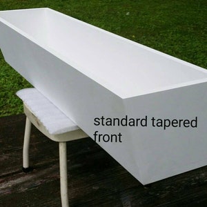 May include: A white rectangular planter box with a standard tapered front. The box is made of wood and is sitting on a small stool. The text "standard tapered front" is visible on the side of the box.