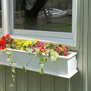 May include: A white window box planter with a variety of colorful flowers, including red, yellow, and green, is mounted on a light green siding wall.