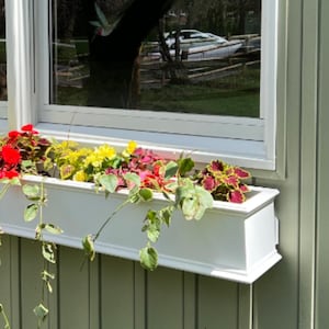 May include: A white window box planter with a variety of colorful flowers, including red, yellow, and green, is mounted on a light green wooden wall.