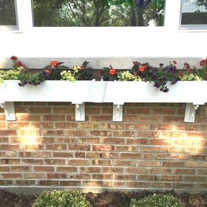 May include: White window box planters with red, orange, and purple flowers. The planters are mounted on a brick wall.