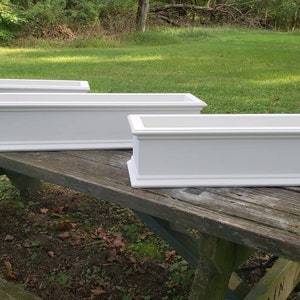 May include: Three white rectangular planters with decorative molding are arranged on a weathered wooden picnic table. The planters are empty and ready for planting. The background features green grass and trees.