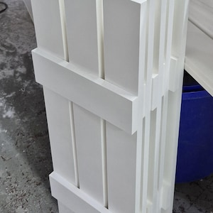 May include: A stack of white wooden shutter panels. Each panel is rectangular with vertical slats and horizontal crossbars. The panels are neatly arranged, showcasing a clean, modern design, likely for home decor or construction.