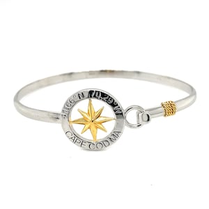 May include: Silver-tone bangle bracelet with a gold compass rose design. The compass rose is centered within a circular frame that reads "CAPE COD MA" and includes coordinates. The clasp is accented with gold-colored wire.